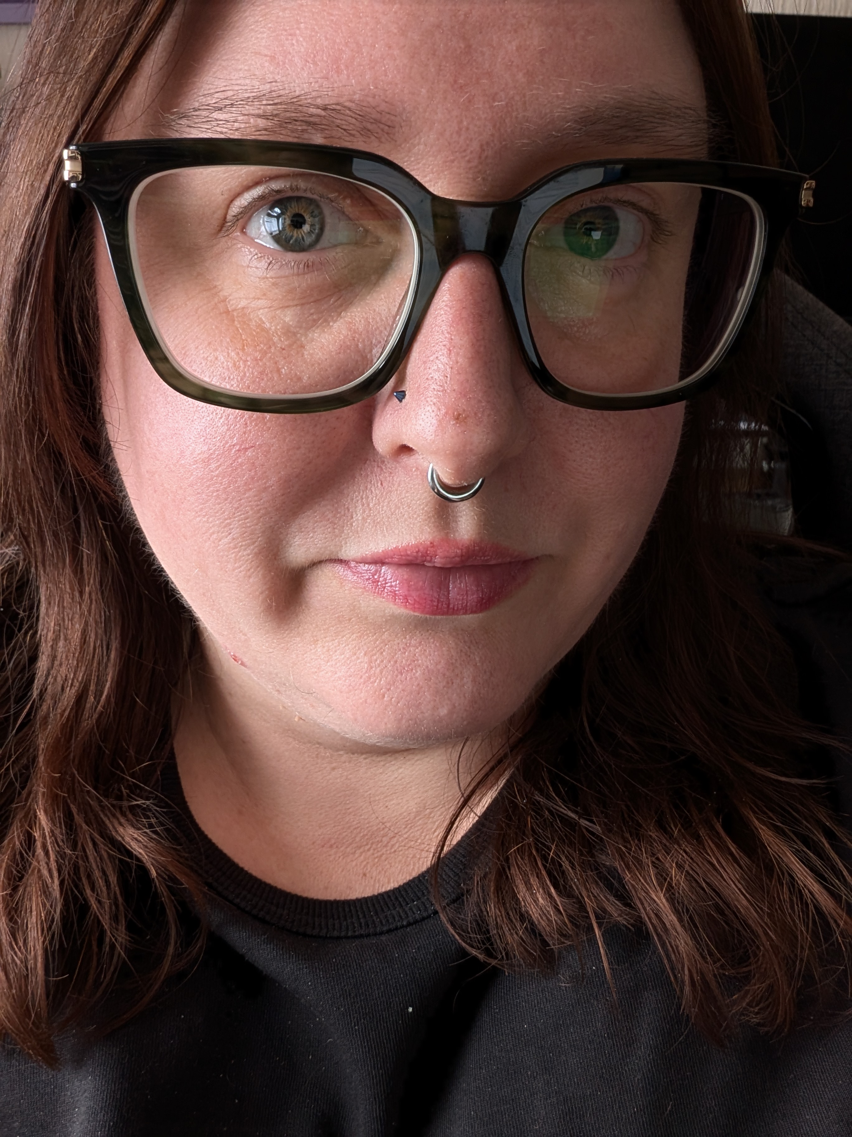 Selfie of a woman with long, dark hair, dark green thick-rimmed glasses and a silver septum ring. She is wearing a black tshirt.